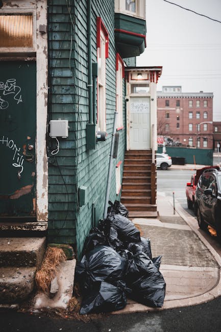 A person wearing a blue and white striped shirt is pushing a black wheeled rubbish bin along a narrow cobbled street between tall brick and stone buildings. The scene is set in an urban environment wi
